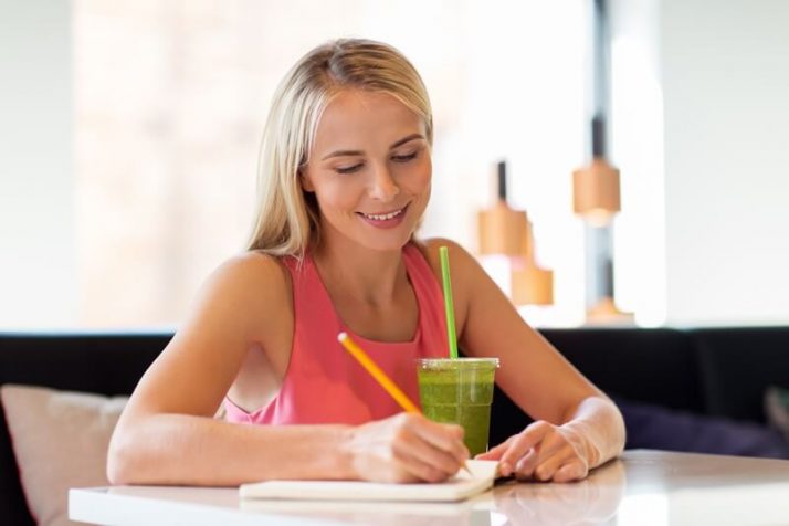 How To Store Smoothies 11 Ways (Fridge, Freezer, How Long); woman with green smoothie drink writing to notebook at restaurant